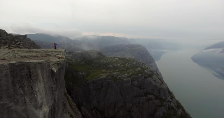 A man and woman stand on a cliff's edge overlooking a vast river valley on a cloudy day