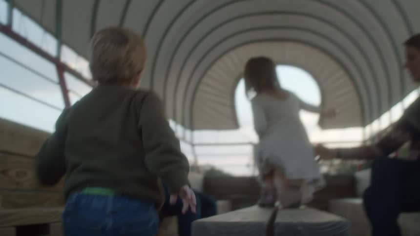 A young boy goes up the stairs to a covered wagon and sits on a bench inside