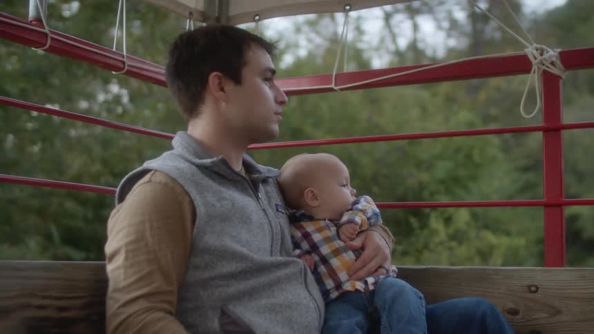 A man in the back of a tractor holds his baby son and smiles