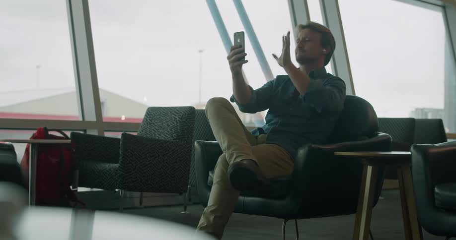 Man sitting in a terminal lounge waves goodbye to a video call on a phone