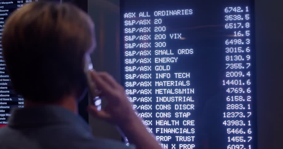 Man talks on a phone in front of a screen with stock info that changes to show other data