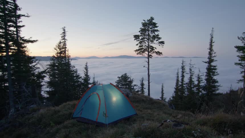 A blue tent sits on a patch of grass overlooking trees and mountains with the sun low in the sky