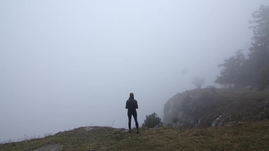 Young fit woman standing on the edge of a cliff in the fog covered mountains