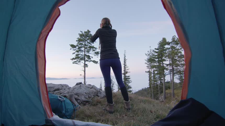 A woman kneeling before the opening of a tent raises to her feet and stretches while looking out at the horizon