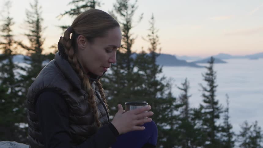 A woman in a forest pouring water into a cup with trees a mountain range the sky and clouds in the background
