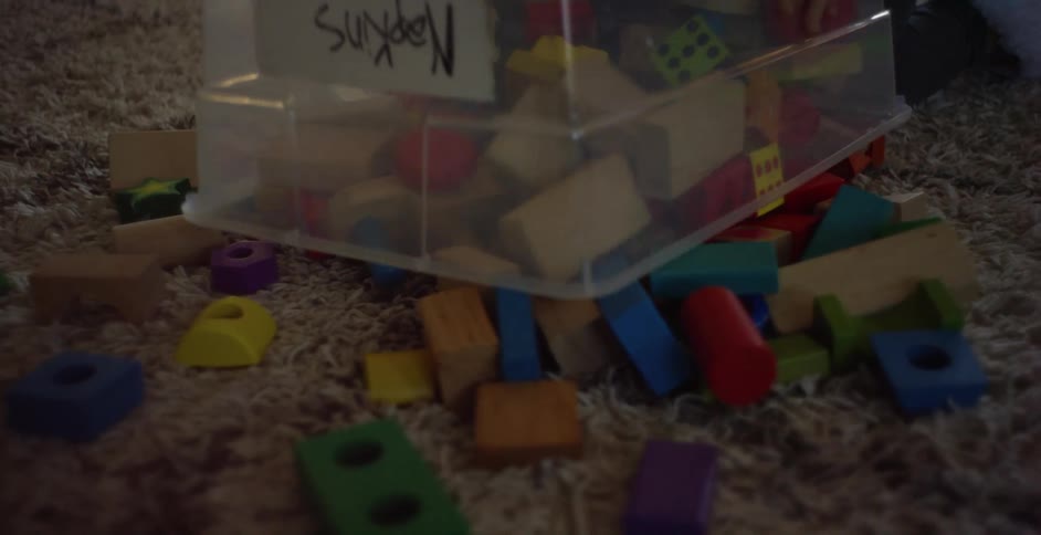 A young girl wearing a pink shirt dumps a bin with toys and blocks onto the family room floor