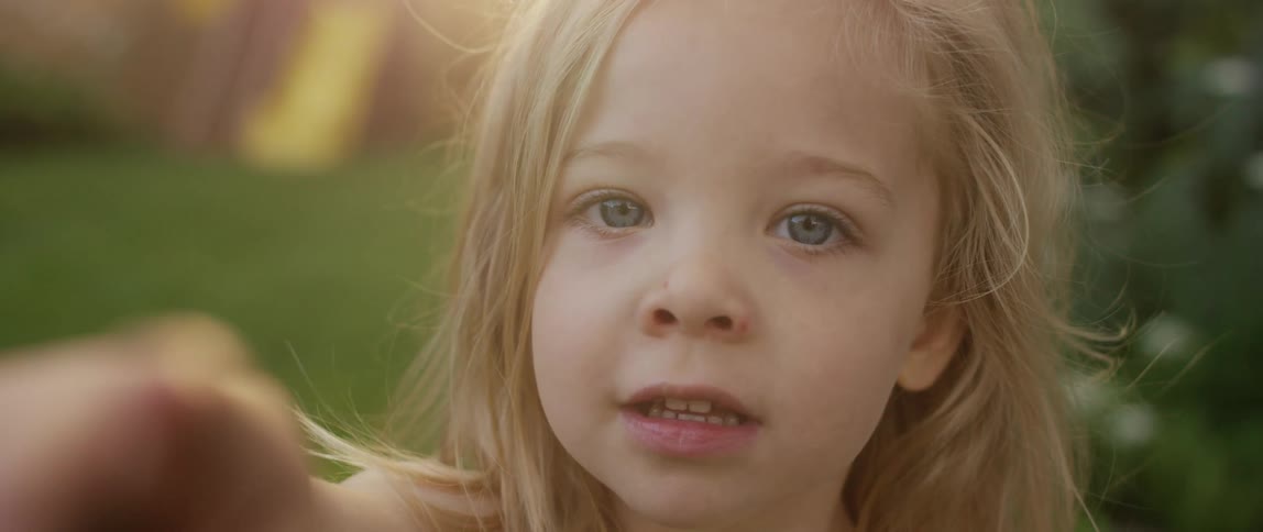 A young girl standing in a fenced yard points into the camera before touching her mouth