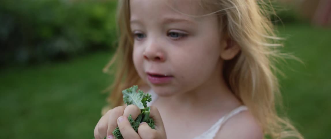 Small blonde girl eating a leaf of lettuce while standing on the grass in the backyard