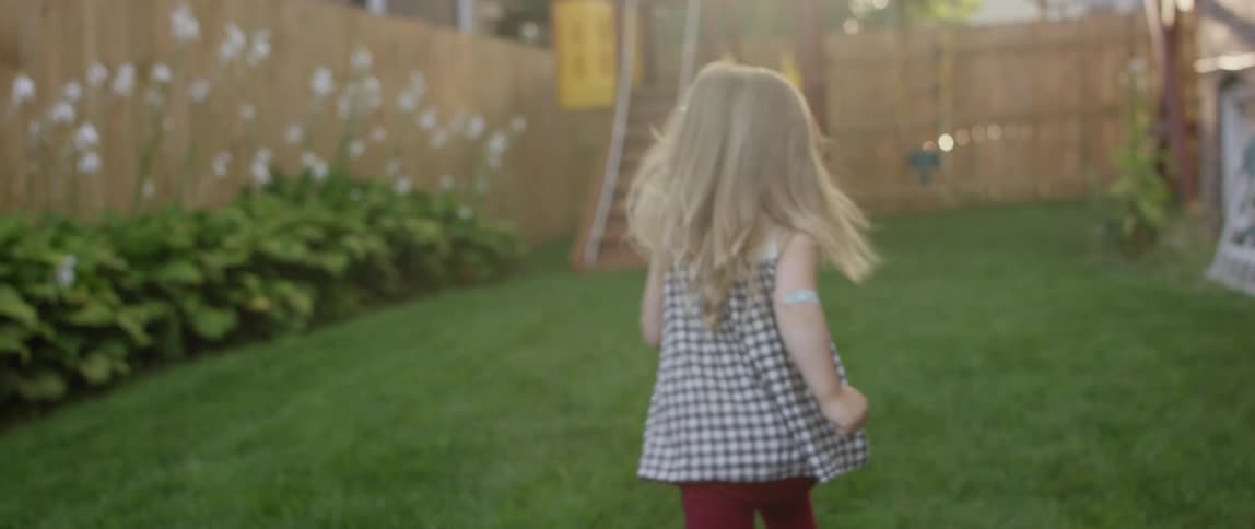 Small girl and a dog run through a fenced in back yard to a swing set on a sunny day