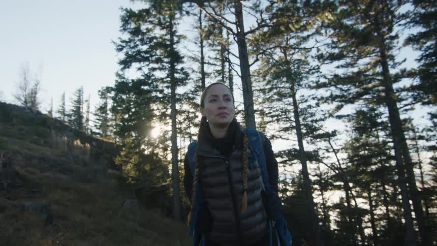 A woman with a backpack in a forest with trees the sky and clouds in the background