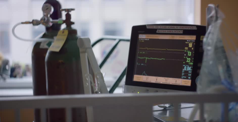 A vital signs monitor next to a bed in a hospital during the day