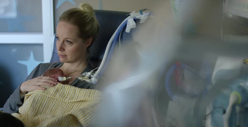 A mother sitting on a chair in the hospital holding her child next to machines