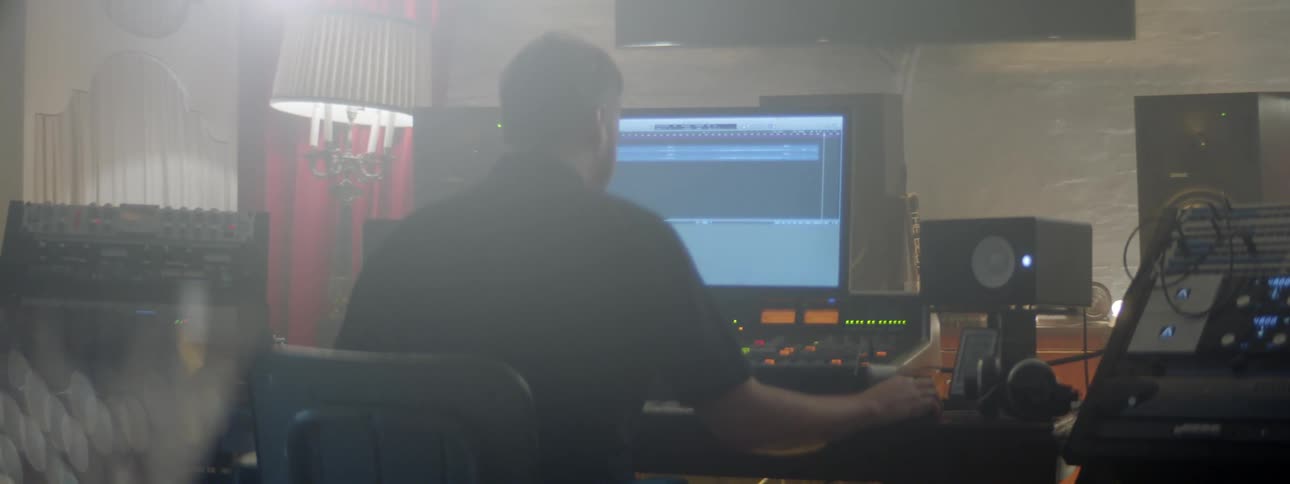 A man sitting on a chair in a music studio in front of equipment with a lamp and a wall in the background