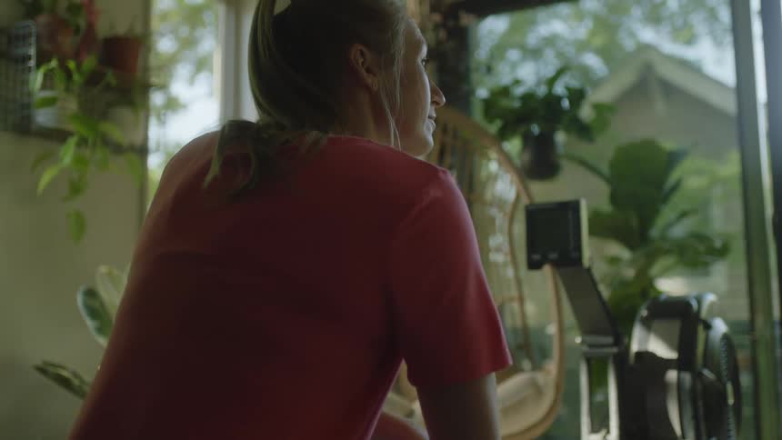 A woman sitting in a room with an exercise machine plants and a glass door in the background