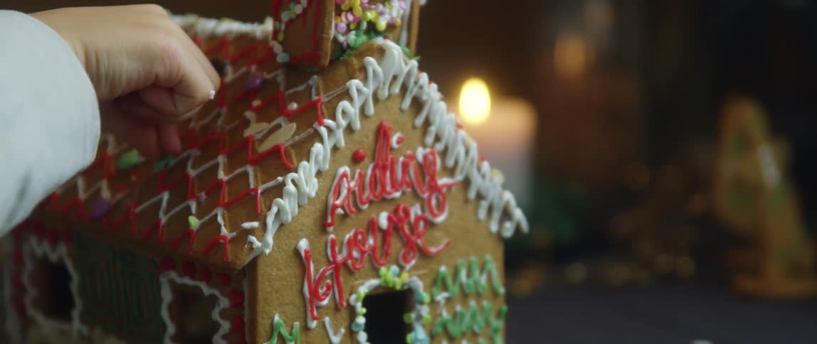 A child places a piece of candy on the roof of a gingerbread house covered in icing