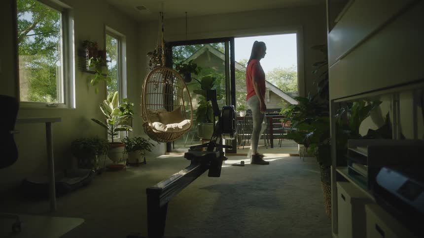 Young woman opens the porch door on the living room and uses a stationary rowing machine