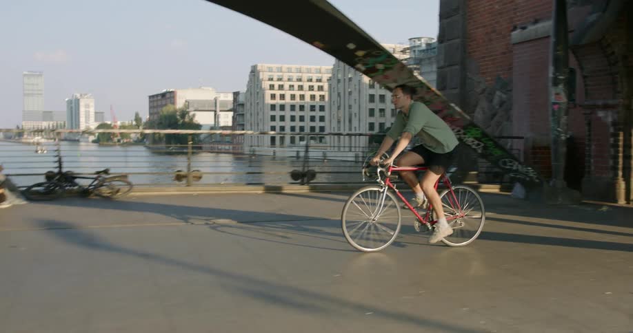 A man wearing shorts and glasses rides a bike on a bridge or boardwalk near a river with buildings in the distance