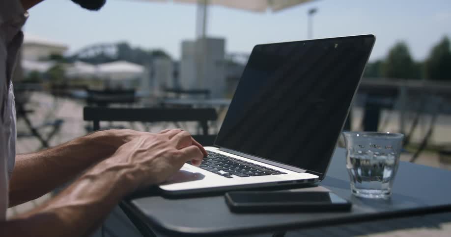Man sits outdoors at cafe working on laptop place on table near mobile phone