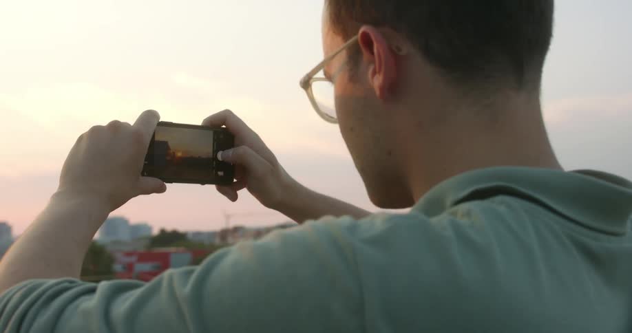 A man with glasses taking pictures on a smart phone with buildings the sky and clouds in the background