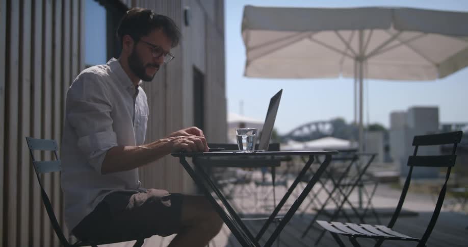 A man sits alone at an outside table at a restaurant and uses his laptop