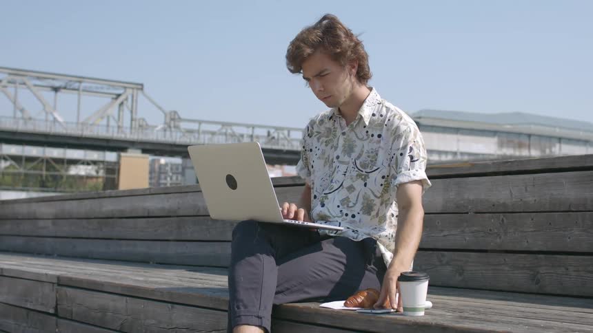 A man sitting on the bleachers next to a croissant typing on a laptop