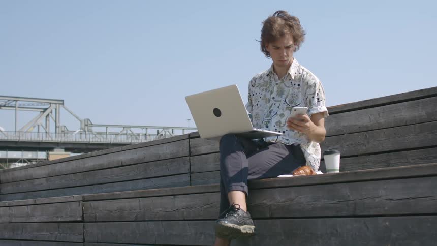 A young man typing on a laptop while sitting outdoors with a croissant and cup of coffee pauses for a moment to answer his smartphone