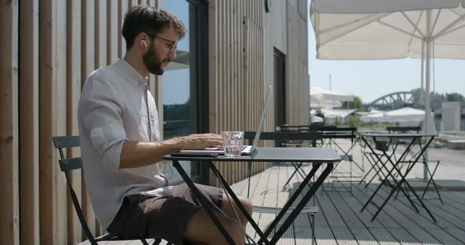 A man wearing shorts sits outdoors at a table working on his laptop and wearing ear buds