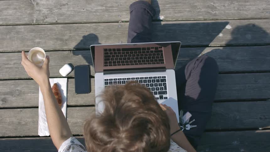A man drinks from a paper cup while sitting outdoors with a laptop on his lap during the day