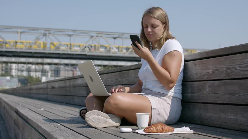A woman sitting on a bench in a city outdoors hangs up her phone and starts typing on her laptop
