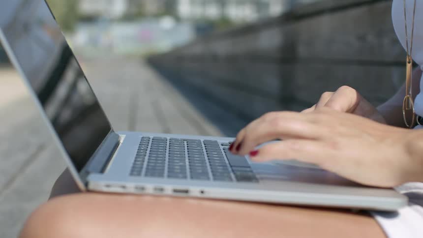 A woman sitting outdoors types on a laptop resting on her legs on a sunny day