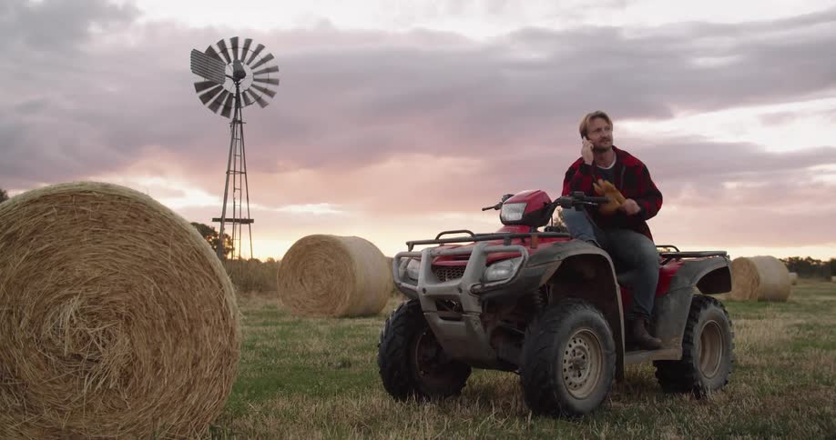 A man talks on his smartphone while sitting on an atv stopped near a bale of hay in a field in the evening
