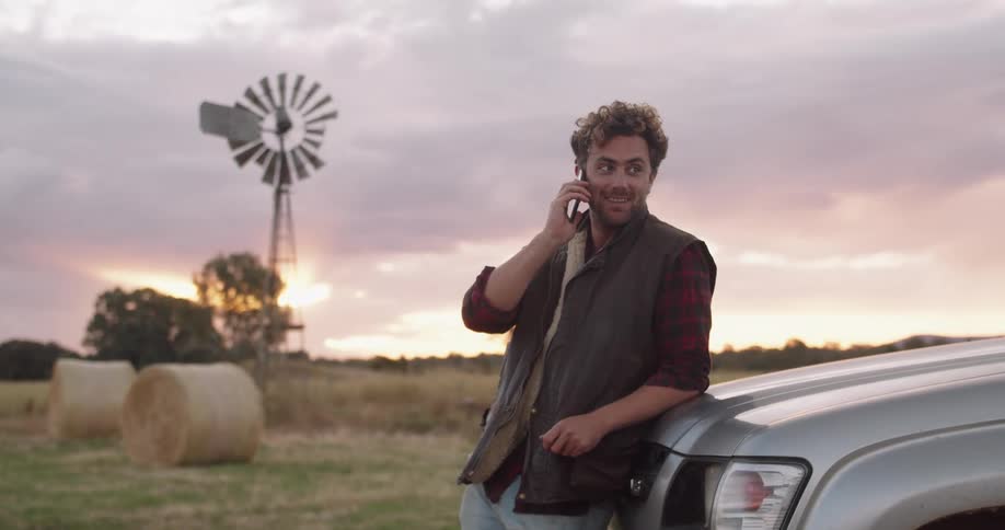 A man on farm land leans against his truck and talks on his cell phone