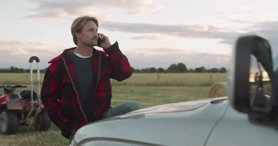 A man talks on a smartphone while standing in a grassland with one foot resting on the bumper of a vehicle