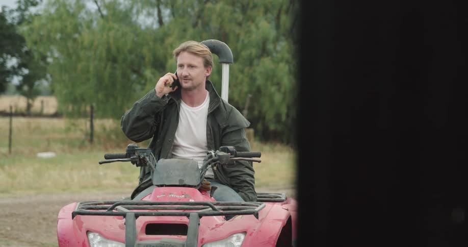 A man sitting on an atv talking on a phone with trees the sky and clouds in the background