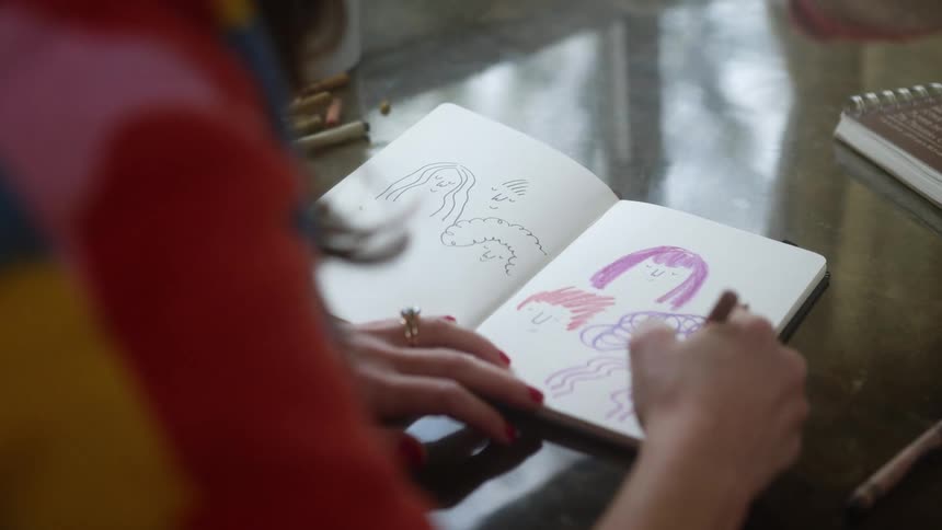 A woman sits at a table at home and draws faces in a notebook with a marker