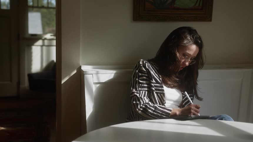 Woman sketches on a table sitting at a table inside of a home dining room at daytime