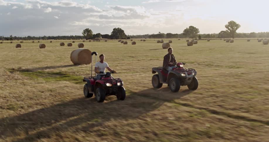 Two men riding four wheelers through a rural field with hay bales