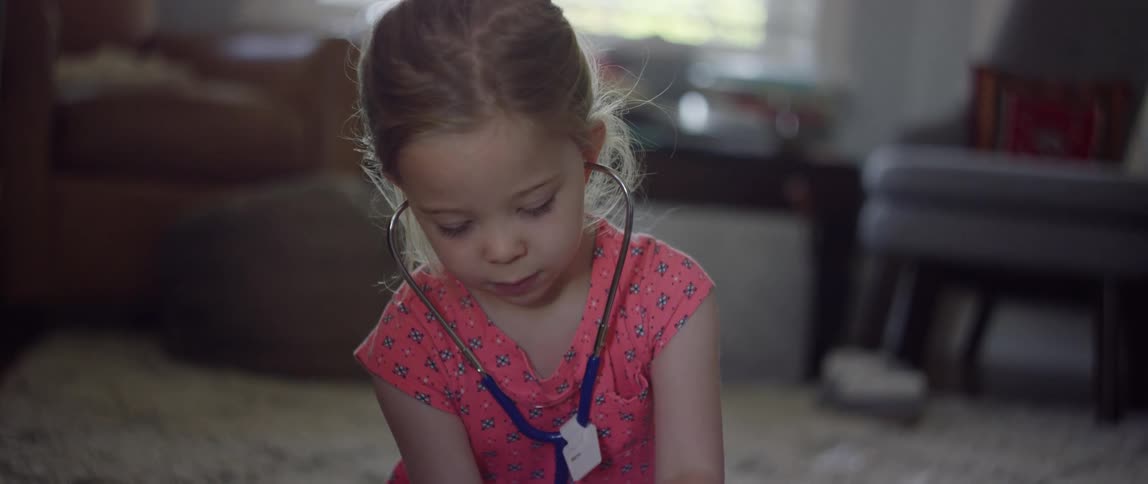 A young girl plays with a stethoscope while sitting on the carpet in the living room of a house during the day