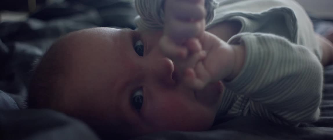 A baby lying on its back atop a bed looks into the camera while fidgeting