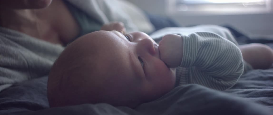 A woman lies next to an infant on a bed and it puts its hands in its mouth