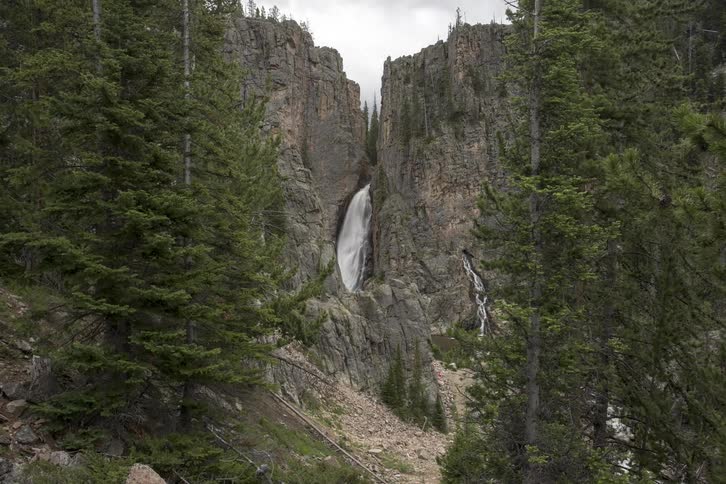 A cliff with a waterfall and river with trees in the foreground and the sky and clouds in the background
