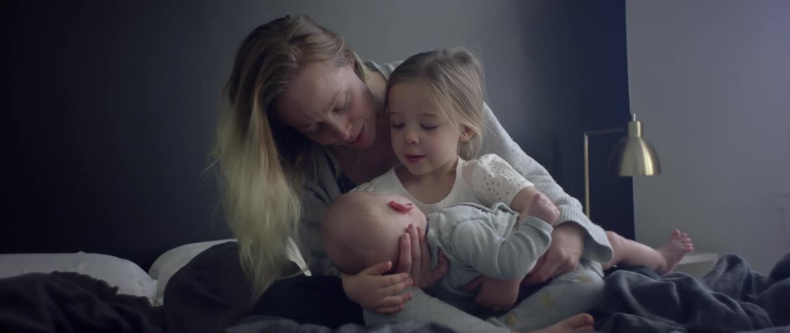 A woman helps her young daughter hold her new sibling as they sit together on a bed