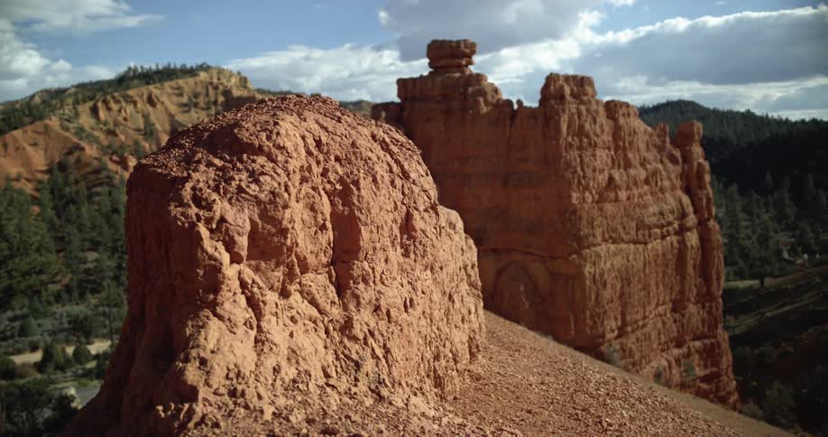 Light red rock formation on the top of a mountain next to other mountains with trees and plants