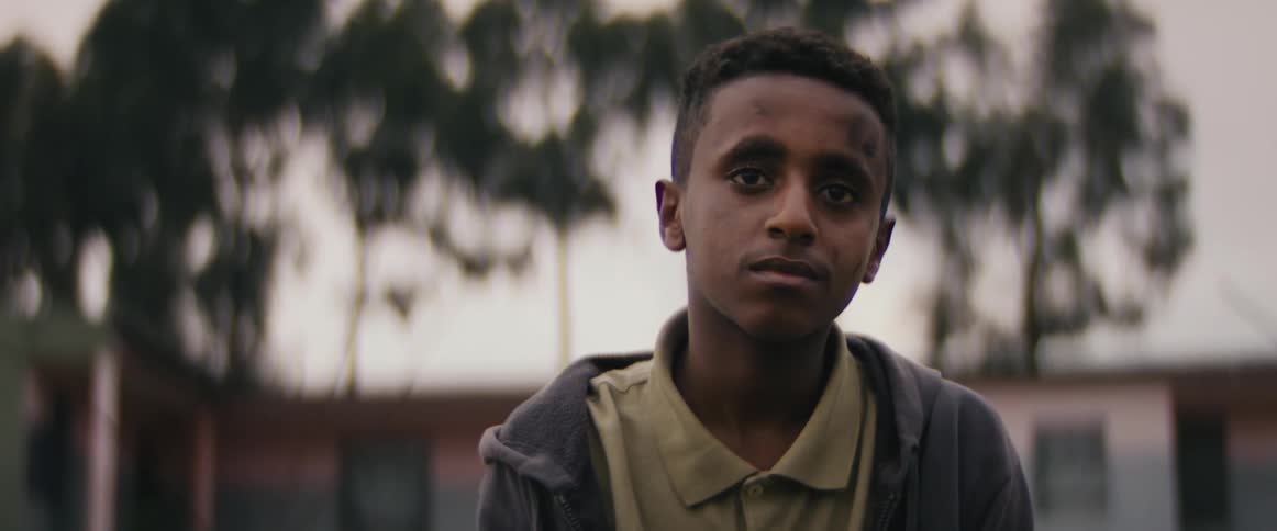 Young boy sits outside in front of a building and stares off into the distance