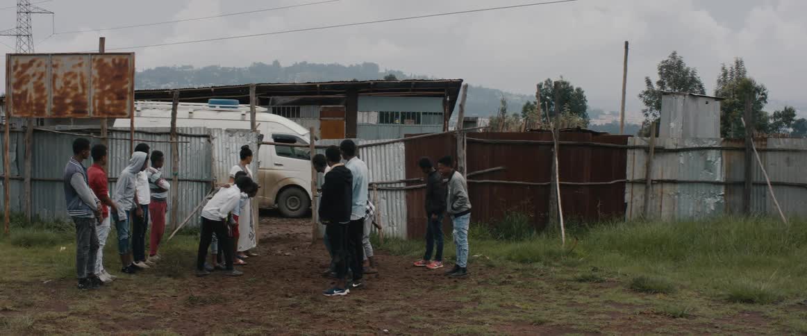 A group of people in a village stand and talk as a van pulls up and more people get out