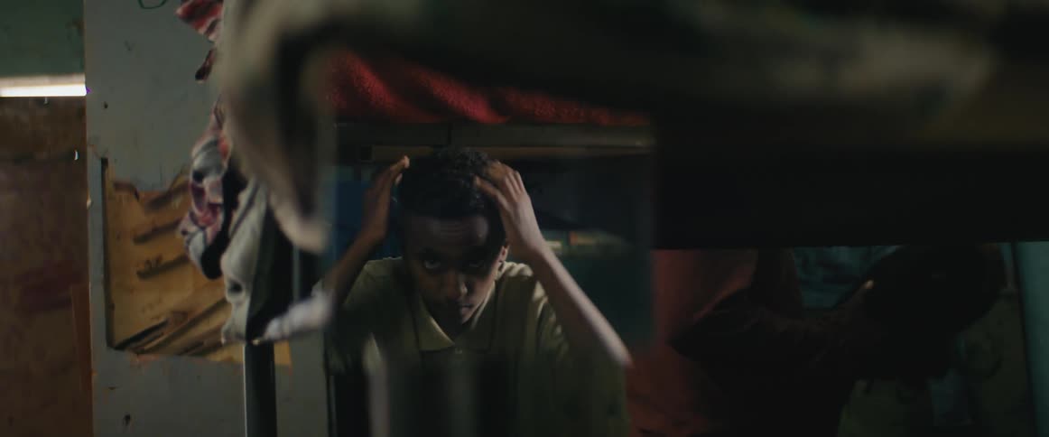 A black male looks in the mirror and adjusts his hair while a person moves around the room behind him