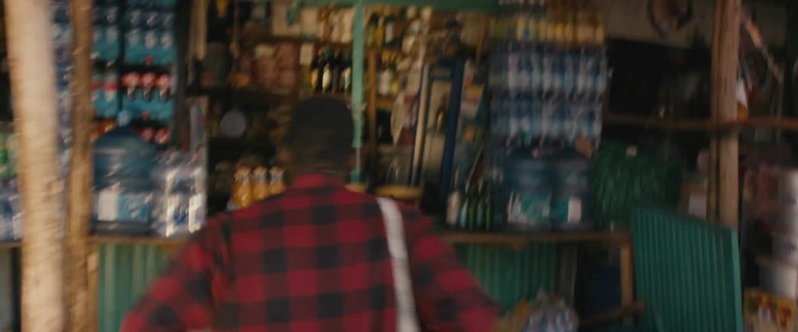 A teenage boy runs in to a shop in a village and steals a drink and runs out to an angry crowd watching him