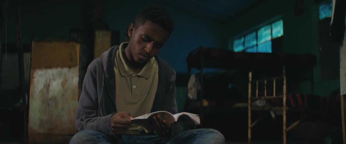 A man in a room sitting and reading a book with beds and windows in the background
