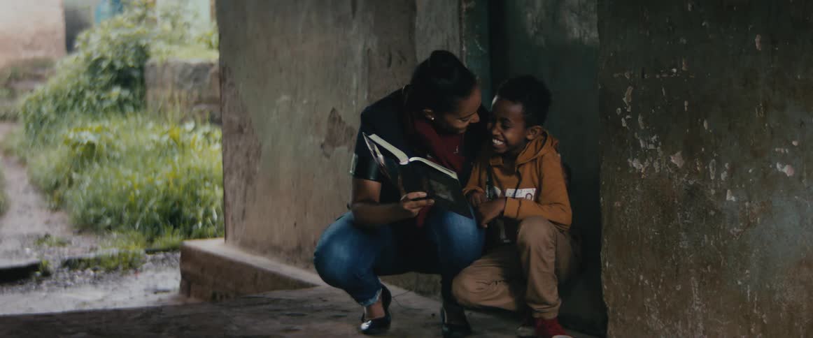 A woman embraces a boy while showing him a book as they kneel together in the doorway of a cement building