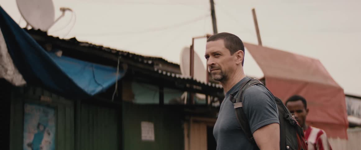 A man holding a piece of paper and wearing a backpack stands in the middle of a village with villagers walking past him